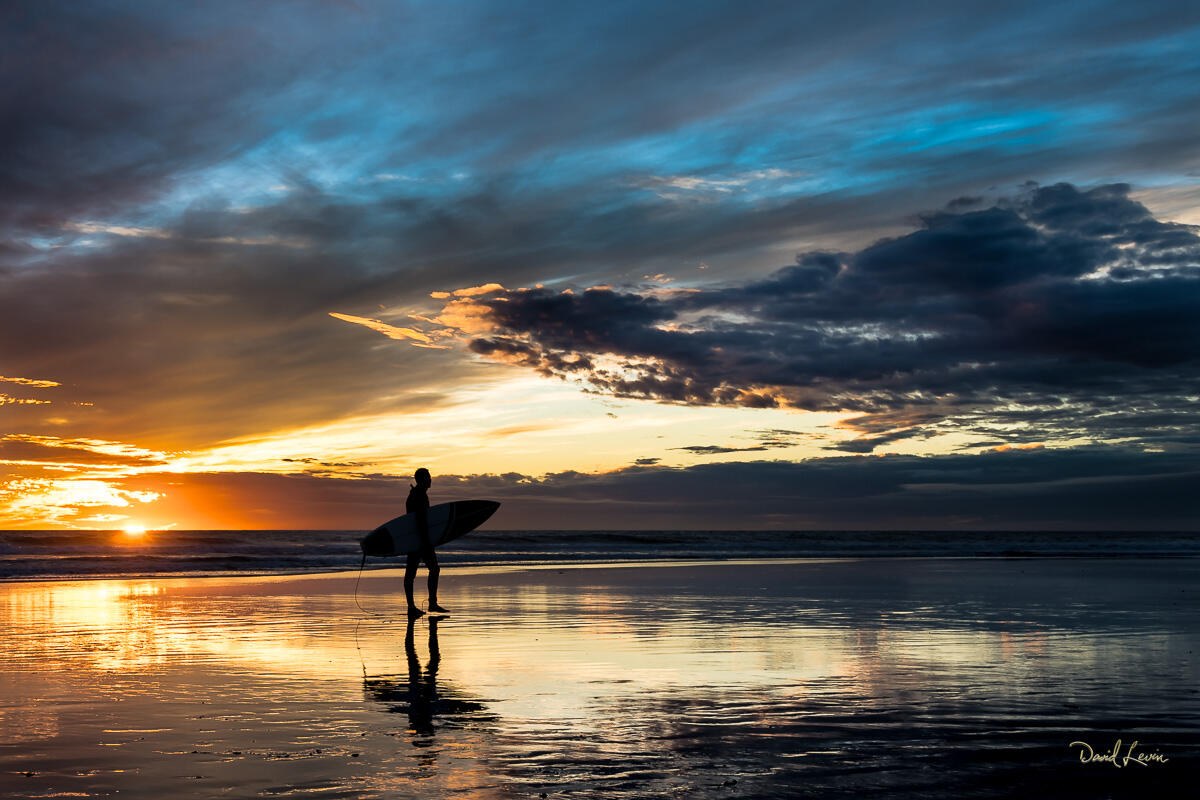 The Last Surfer Lone surfer walking along an Oceanside beach at sunset with warm coastal light.
