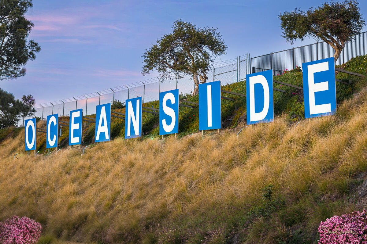 Oceanside Sign Oceanside sign on a coastal hillside with blue letters, grasses, flowers, palm trees, and soft evening light.